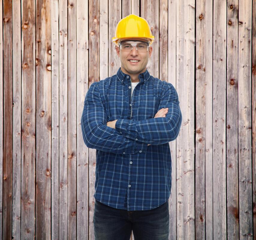 A picture of a worker standing in front of a fence with arm crossed and hard hat on
